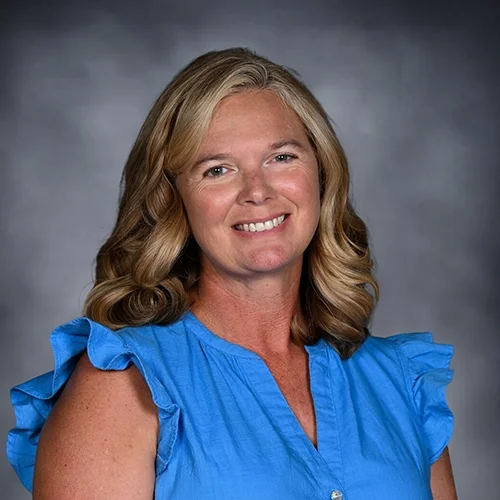 Smiling woman with blonde hair wearing a blue and white patterned blouse, posed against a gray background.