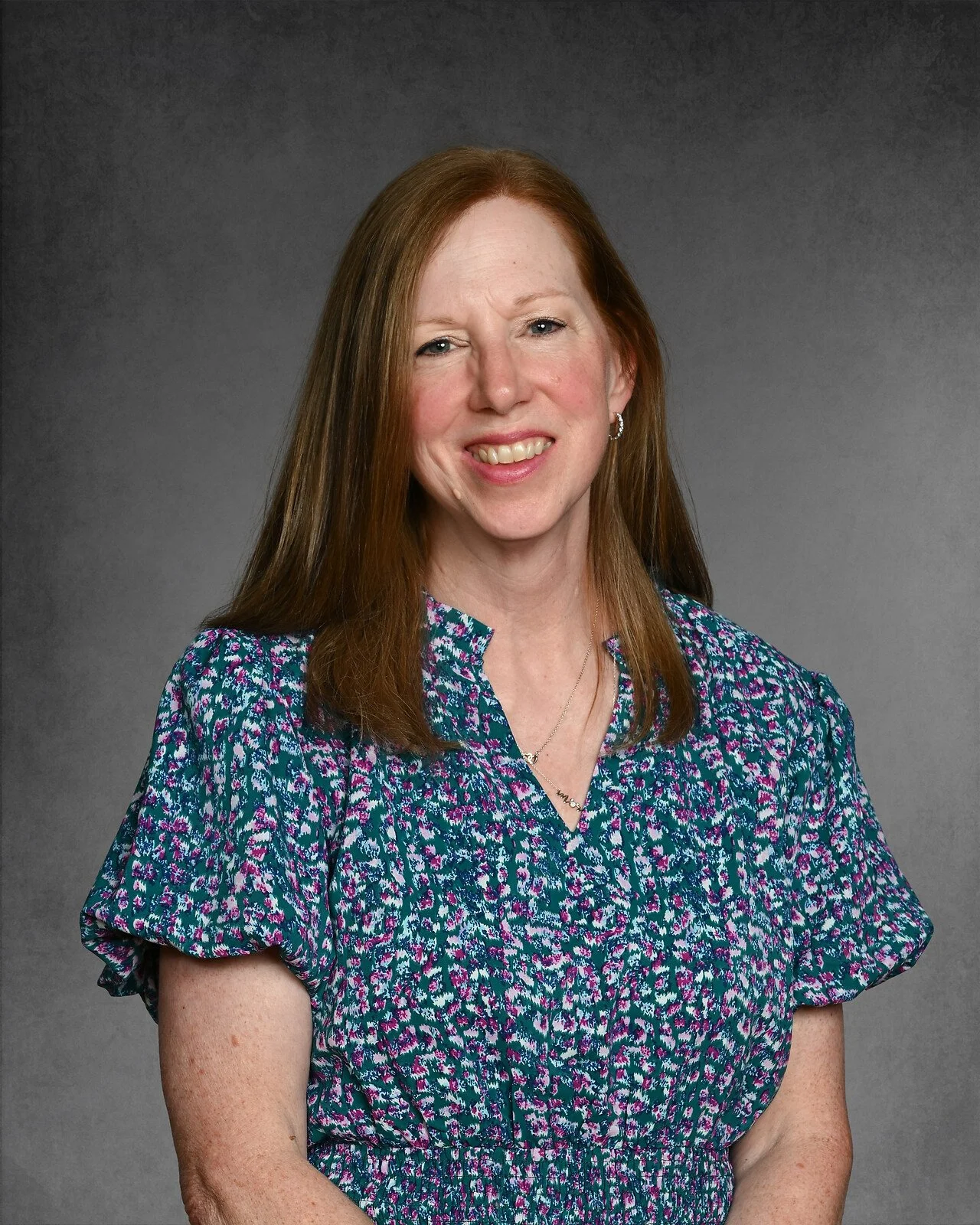 A woman with shoulder-length hair wearing a polka dot blouse, smiling against a gray background.