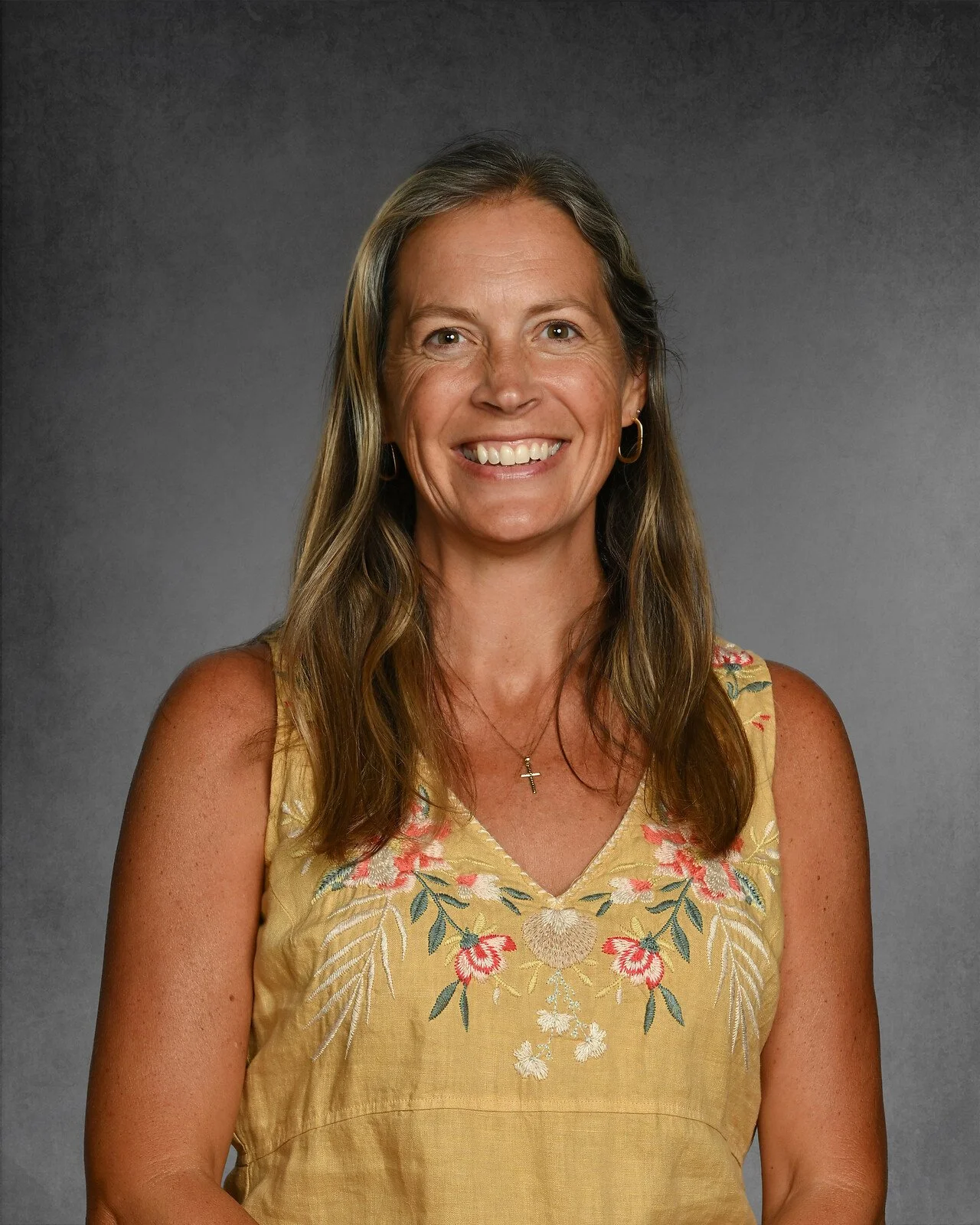 Smiling woman with long hair wearing a floral top and hoop earrings against a gray background.