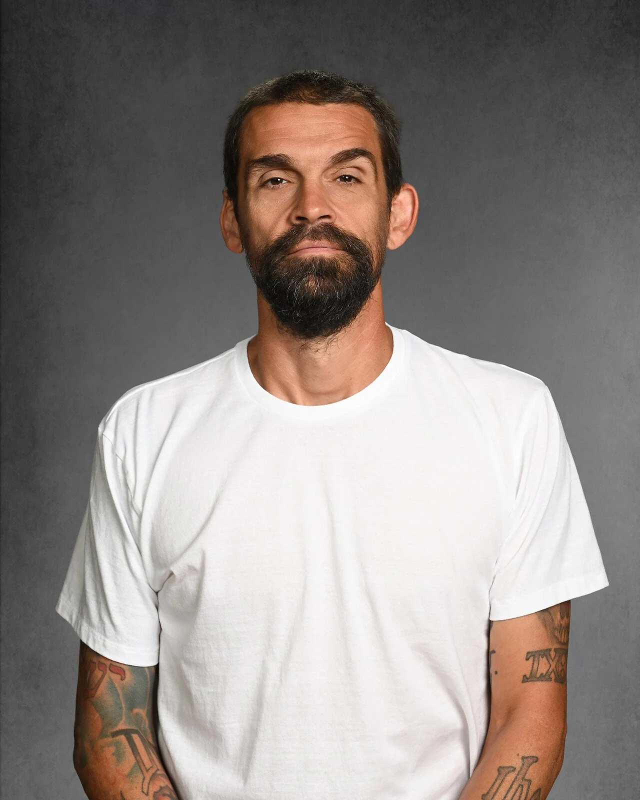 A man with long dark hair, a beard, wearing a black shirt and black cap, standing against a dark gray background.