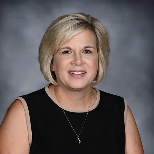 Woman with short blond hair wearing a black top and necklace, smiling against a gray background.