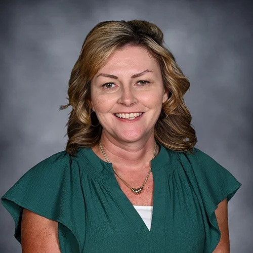 Woman with blonde hair wearing a floral-patterned blouse, smiling against a gray background.