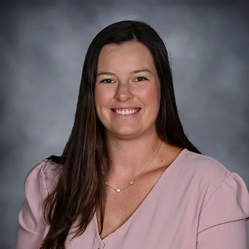 Smiling woman with long dark hair wearing a colorful plaid dress against a gradient background.