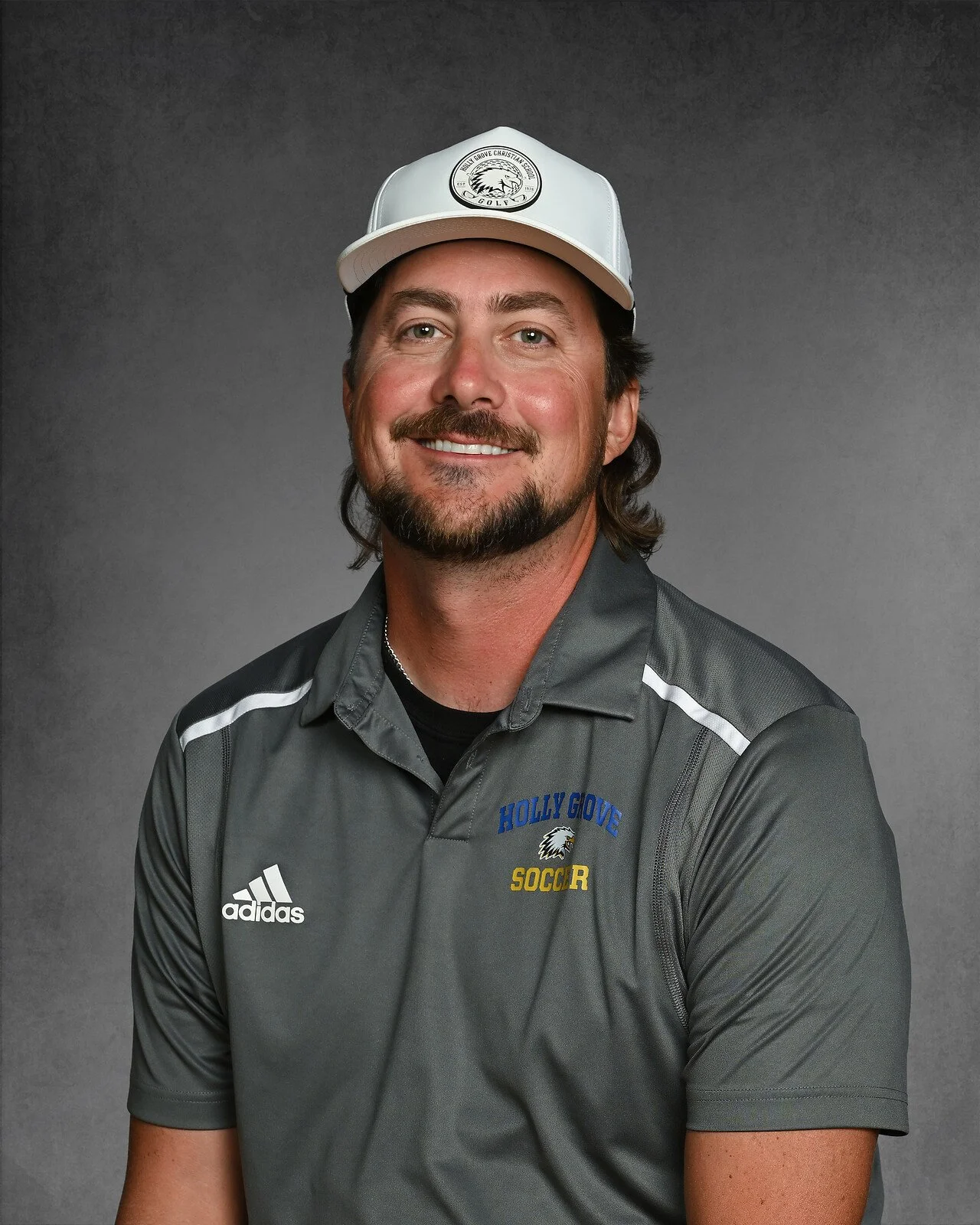 A man with dark hair and a beard wearing a charcoal gray polo shirt with the logo of Holly Grove Christian School Athletics, smiling against a dark gray background.