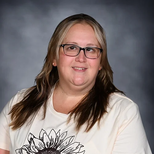 Portrait of a woman with blonde and dark brown hair, wearing glasses and a green sweater, against a gray background.