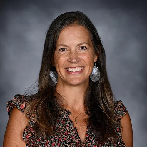 Smiling woman with long hair wearing a floral top and hoop earrings against a gray background.