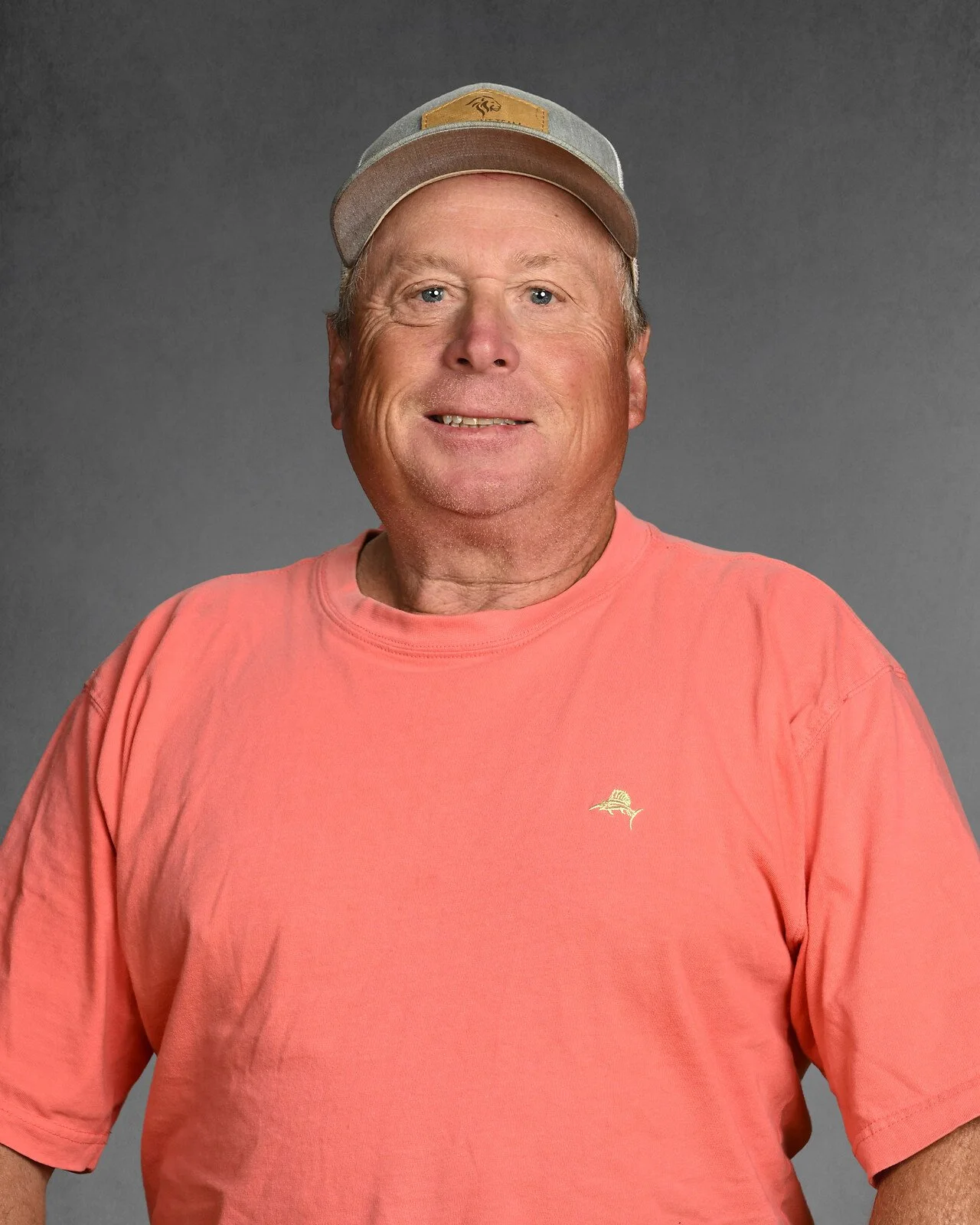 A smiling man wearing a gray baseball cap and a light blue T-shirt against a gray background.