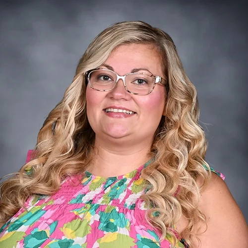 A woman with long blonde hair, wearing glasses and a floral blouse, smiling at the camera against a gray background.