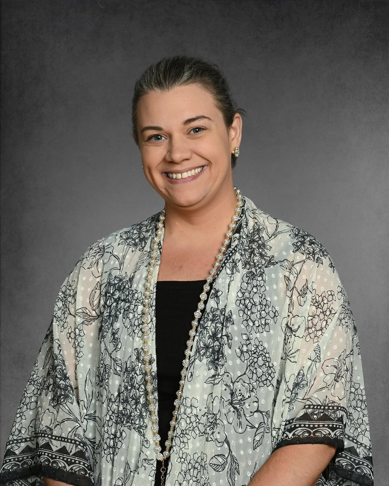 A woman with shoulder-length wavy blonde hair and bangs, smiling, wearing a black top and a necklace with a small pendant, against a gray background.