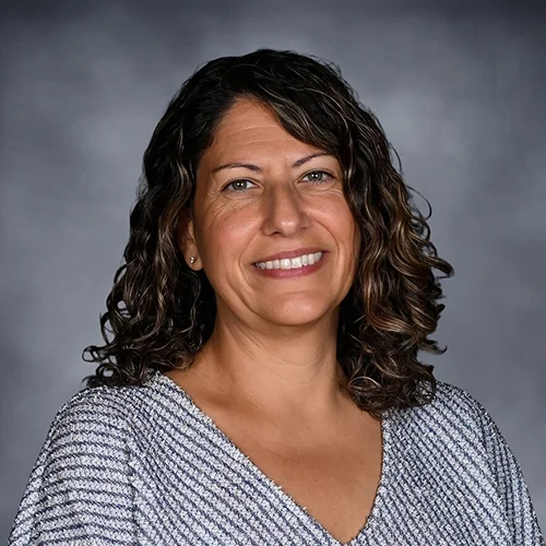 Smiling woman with curly hair wearing a floral blouse against a gray background.