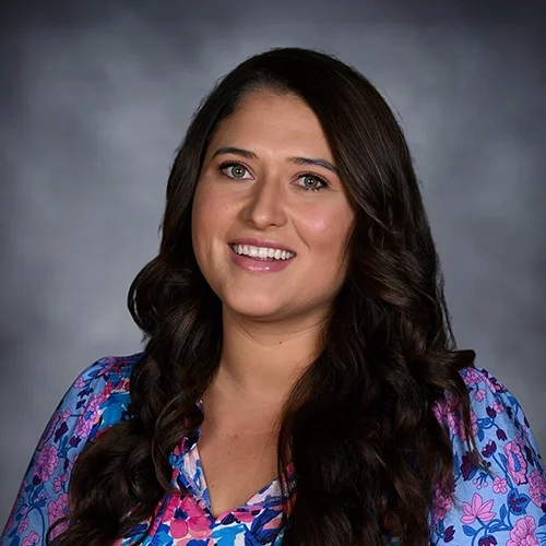 Woman smiling in a colorful shirt with long brown hair against a gray background.