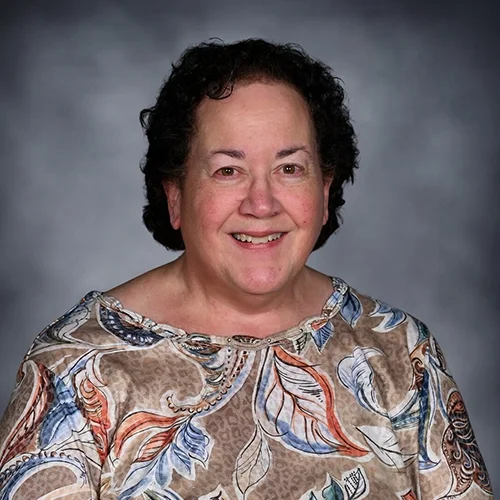 Smiling woman with glasses and earrings wearing a red patterned blouse, set against a gray background.