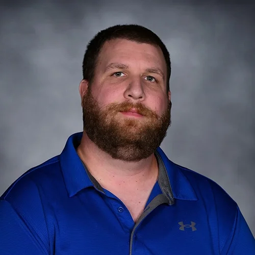 A man with short hair, a beard, and a mustache smiling, wearing a dark patterned shirt, against a plain gray background.