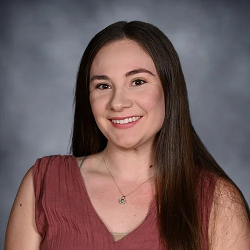 A young woman with long brown hair, smiling, wearing a floral sleeveless top and a necklace against a dark background.