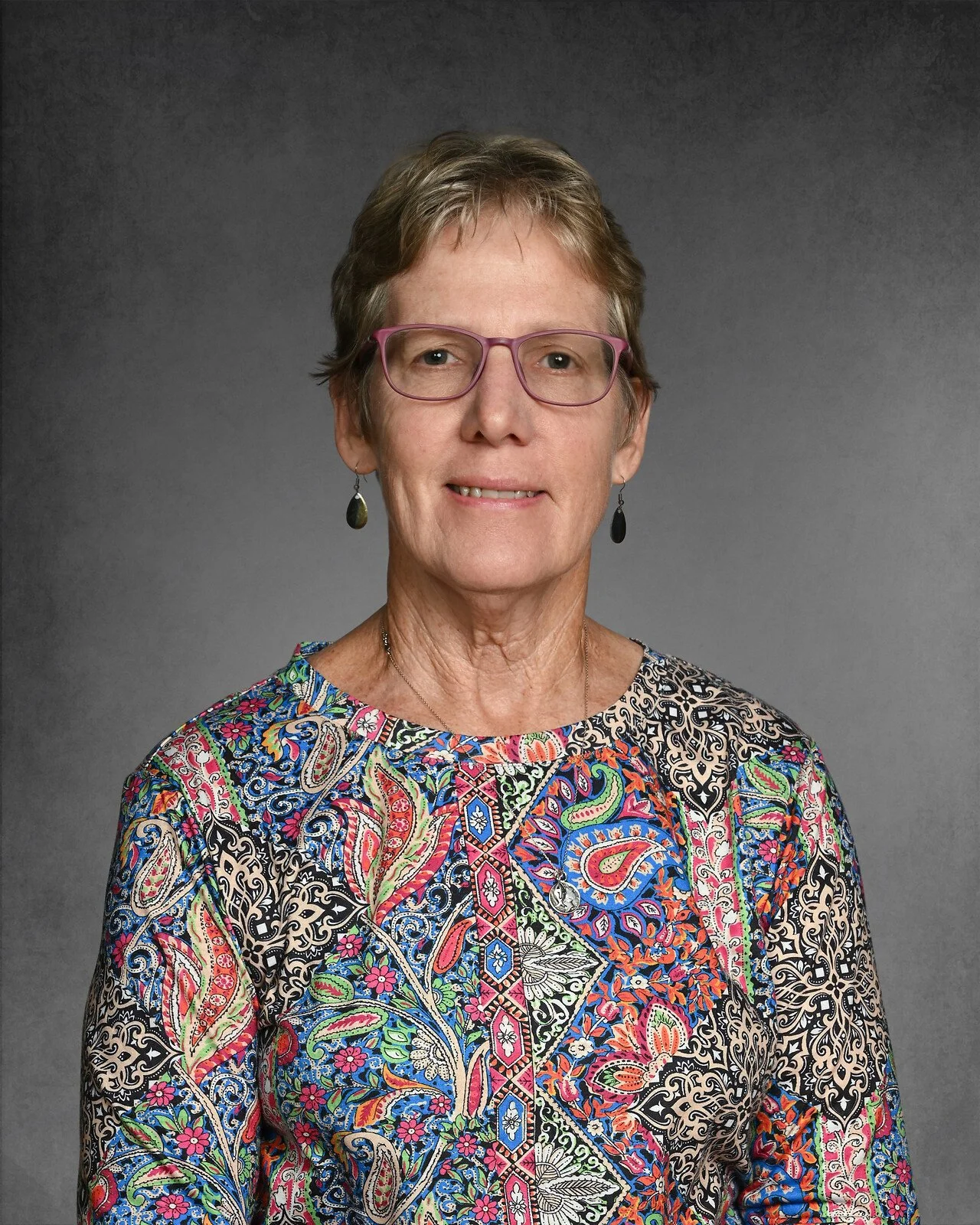 A smiling elderly woman with short hair wearing glasses, pink earrings, and a vibrant pink and orange patterned top.
