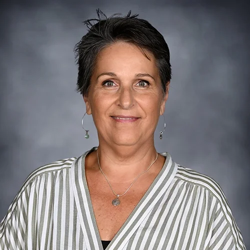 Smiling woman with short dark hair, wearing a white blouse and a patterned vest, with earrings, against a gray background.