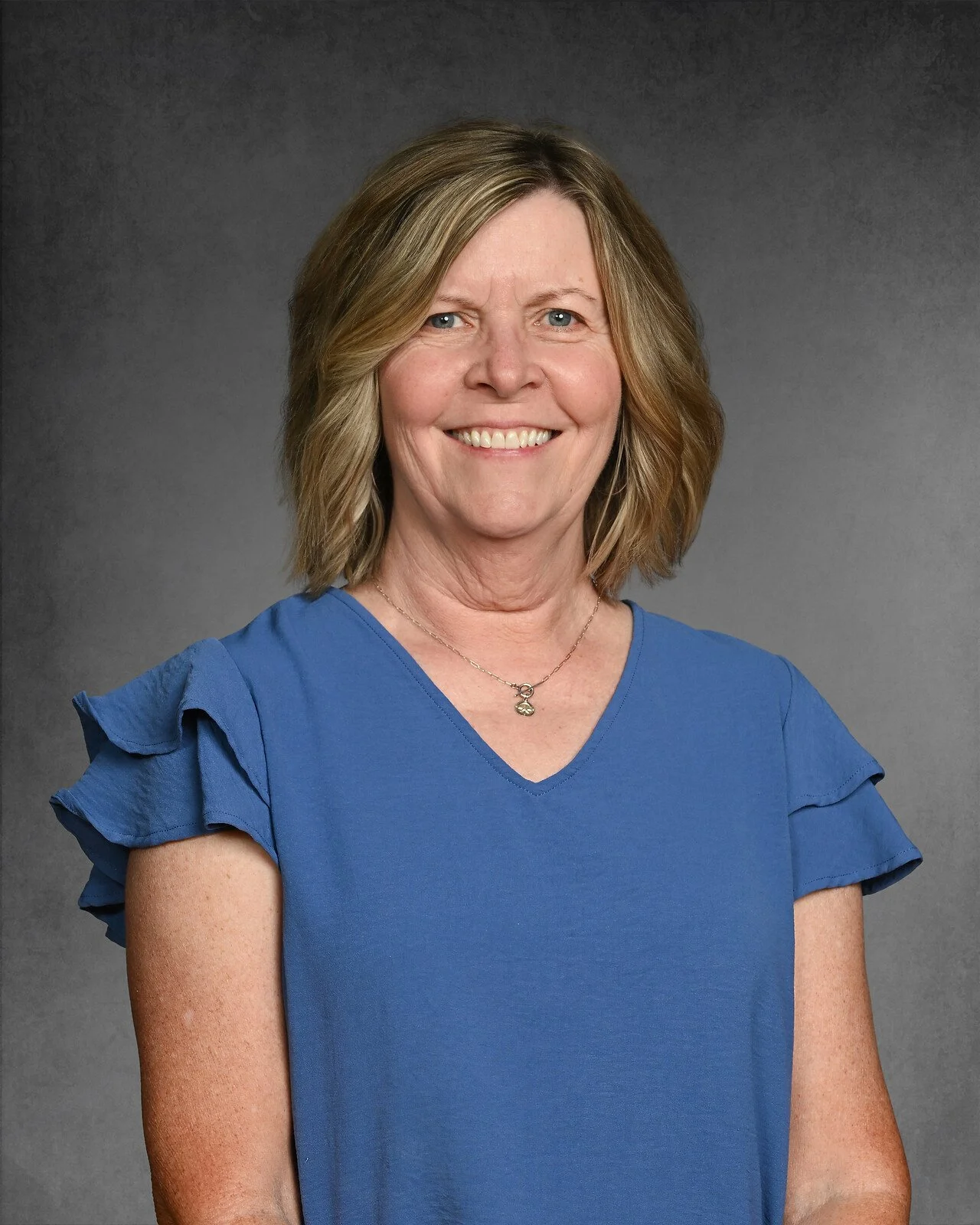 A woman with shoulder-length light brown hair smiling, wearing a blue blouse, against a dark gray background.