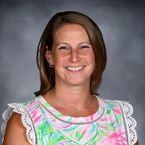 Smiling person with short hair wearing a teal blouse and dark earrings, against a gray background.