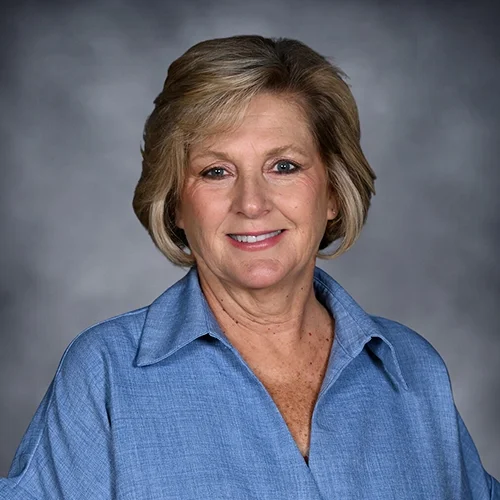 A smiling middle-aged woman with blonde hair in a navy blue dress, posing against a gray background.