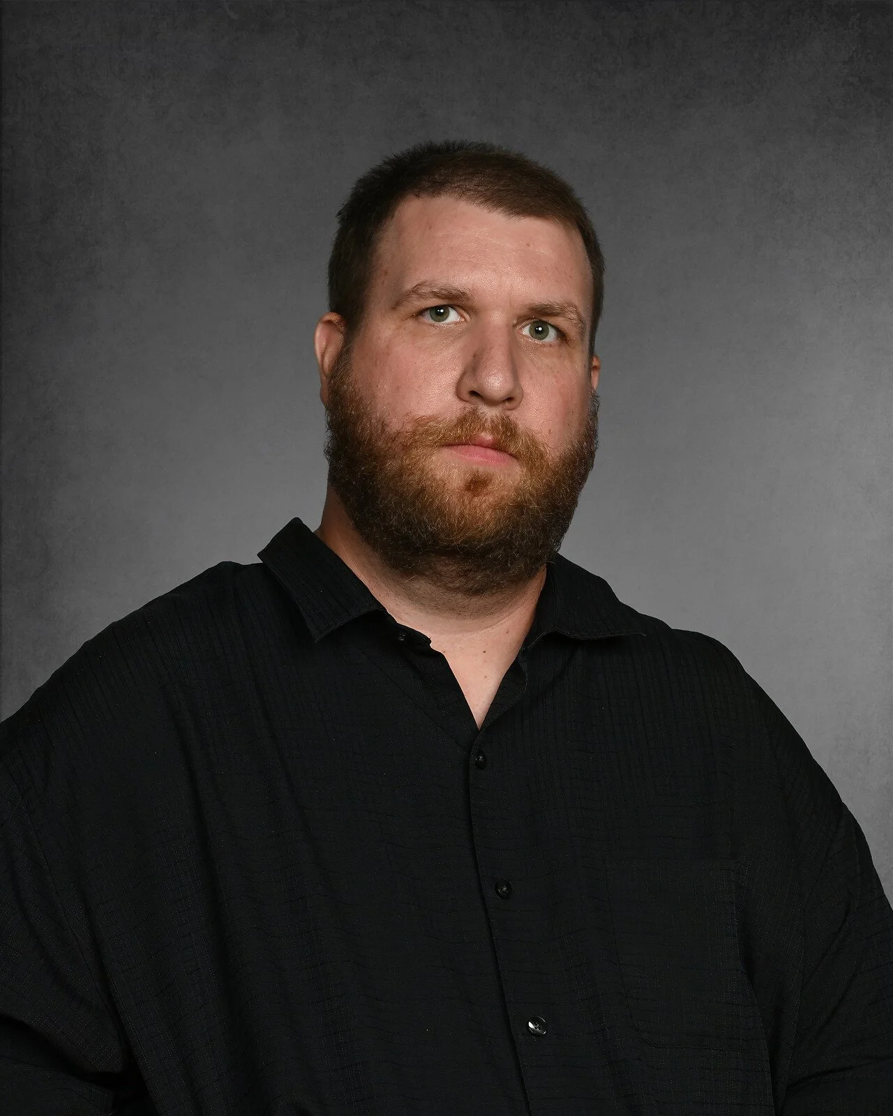 A man with short hair, a beard, and a mustache smiling, wearing a dark patterned shirt, against a plain gray background.