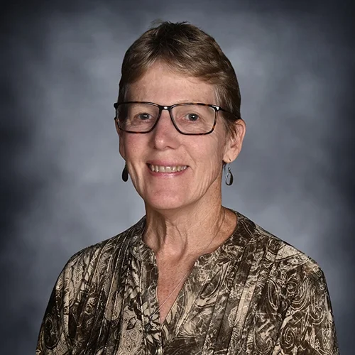 A smiling elderly woman with short hair wearing glasses, pink earrings, and a vibrant pink and orange patterned top.