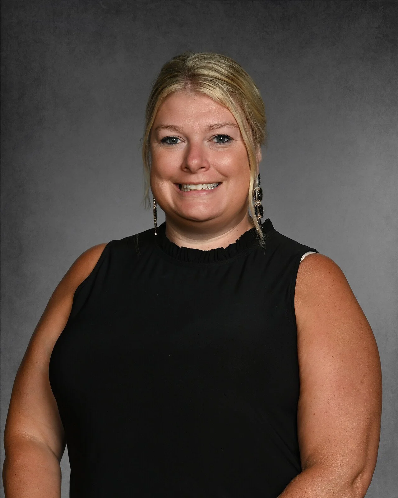 A woman with blonde hair, smiling, wearing large pearl earrings and a sleeveless black top, posing against a gray background.