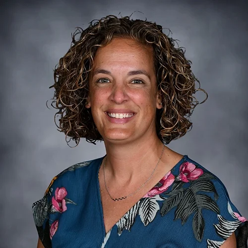 A woman with curly hair smiling, wearing a white sleeveless top with gray and black stripes, standing against a gray background.