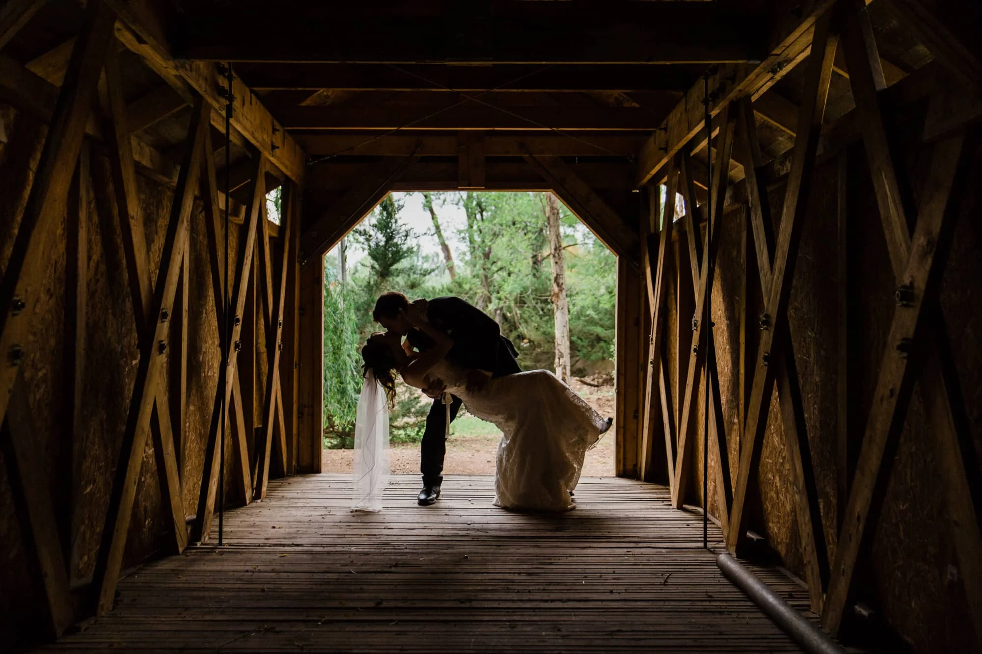 Groom dipping bride and kissing under a covered bridge, silhouetted against bright outside light. 