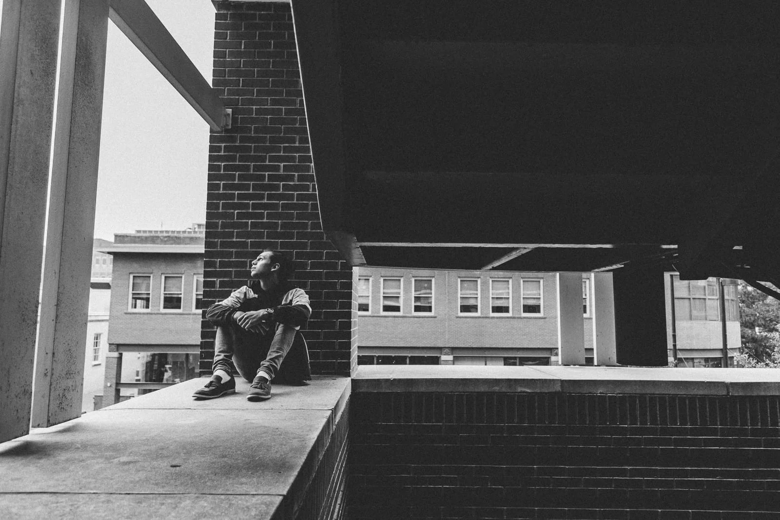 Wide portrait of a young man sitting on the ledge of a stair well and looking up, in black and white.