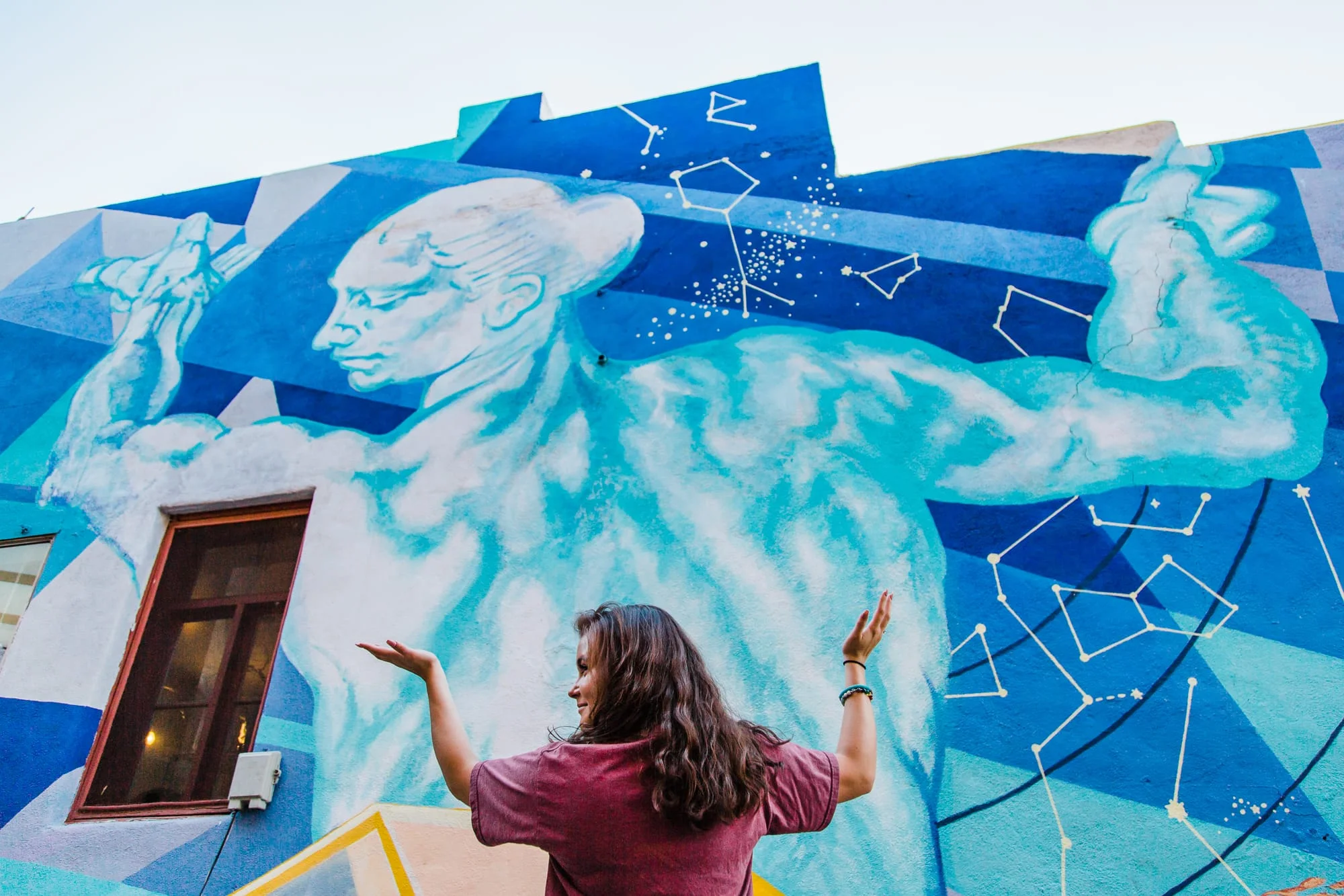 Wide portrait of young woman standing in front of a mural of Atlas and mimicking his pose.