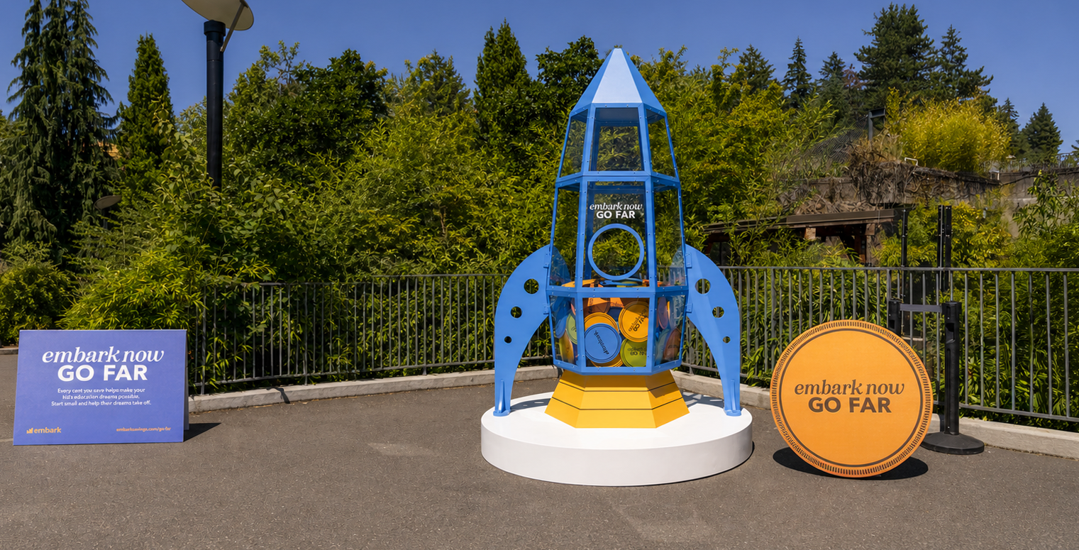 Embark Now, Go Far rocket-shaped piggy bank with a branded sign and oversized coin at the Oregon Zoo.