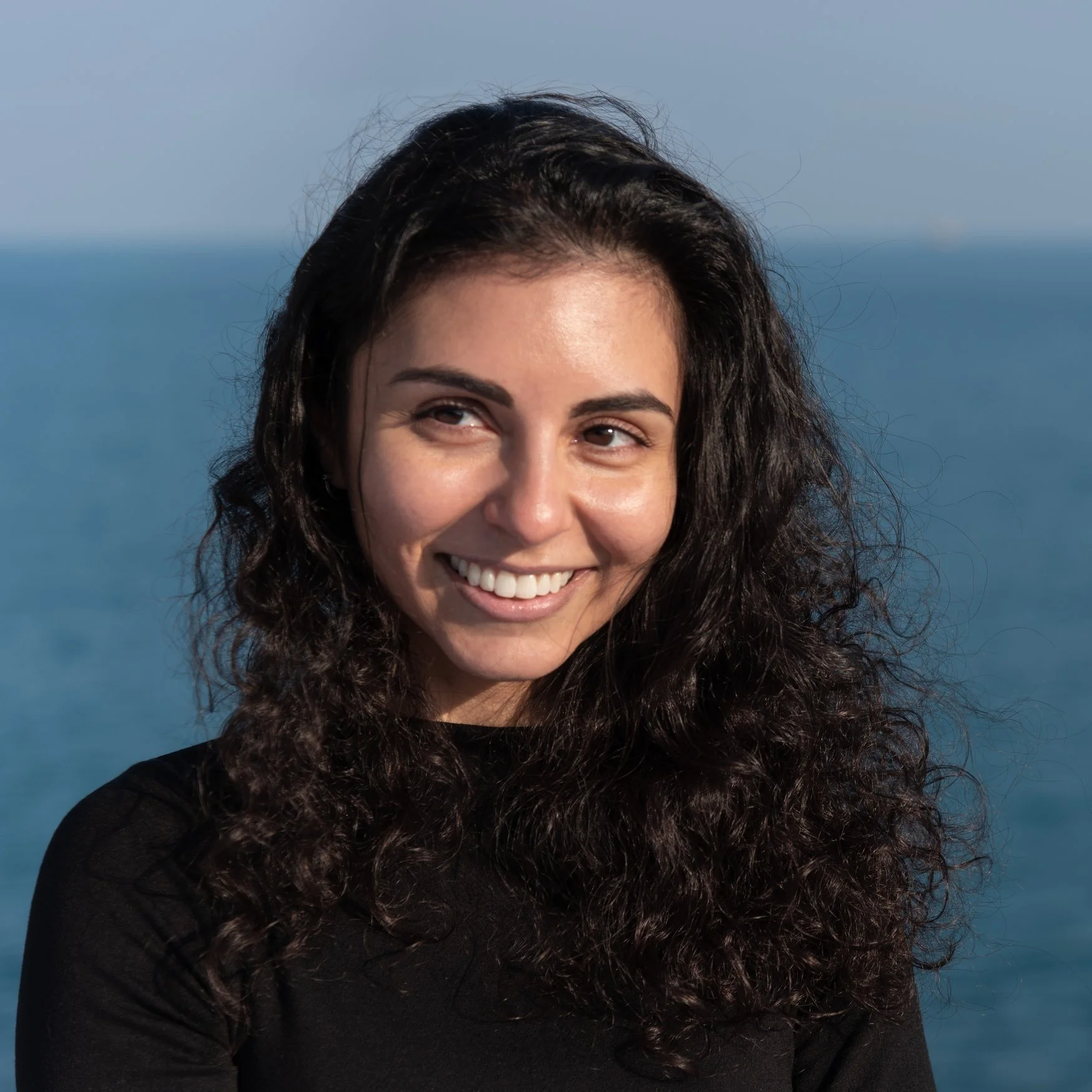 Woman with light brown skin and dark curly hair wearing a black sweater. She is smiling while looking off to the left. Behind her is Lake Michigan and her hair is blowing in the wind.