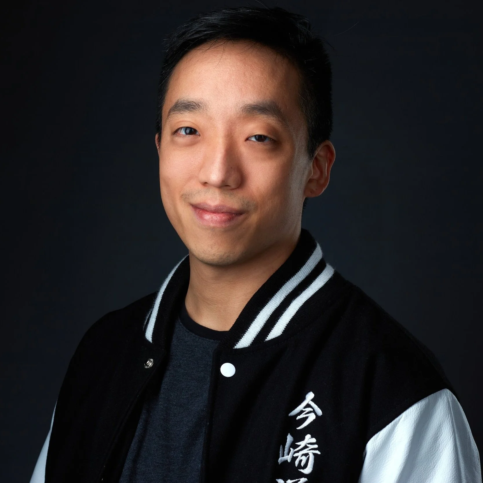 Vietnamese-American man smiles, closed mouthed, at the camera. He is wearing a dark black shirt and a black letterman-style jacket with white stripes on the collar