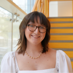 A smiling white person with shoulder-length brown hair with bangs and glasses, wearing a white blouse and beaded necklace.