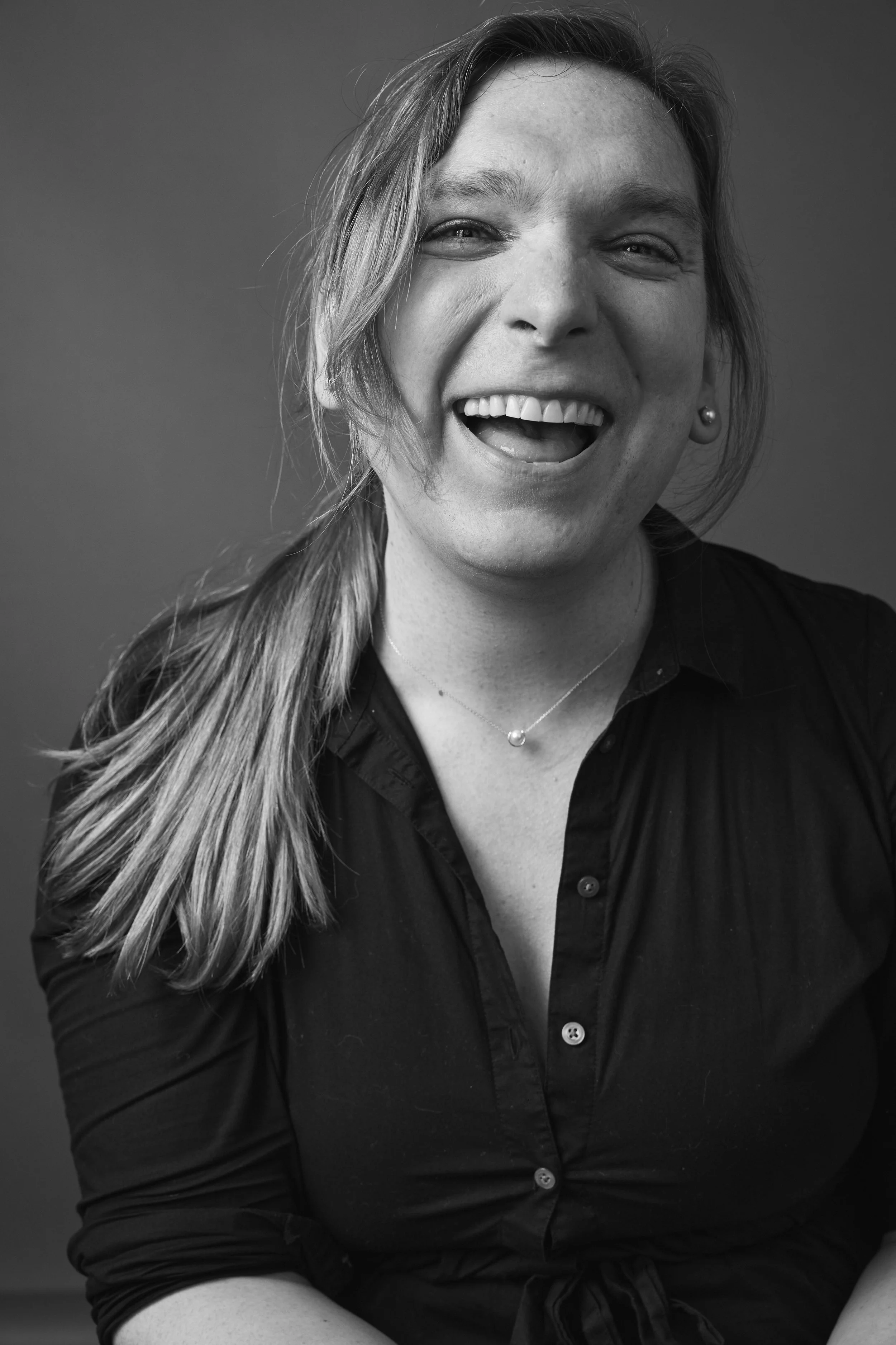 A white woman smiling broadly at the camera, with long hair pulled back, wearing a dark button-up shirt and necklace in a black-and-white portrait.