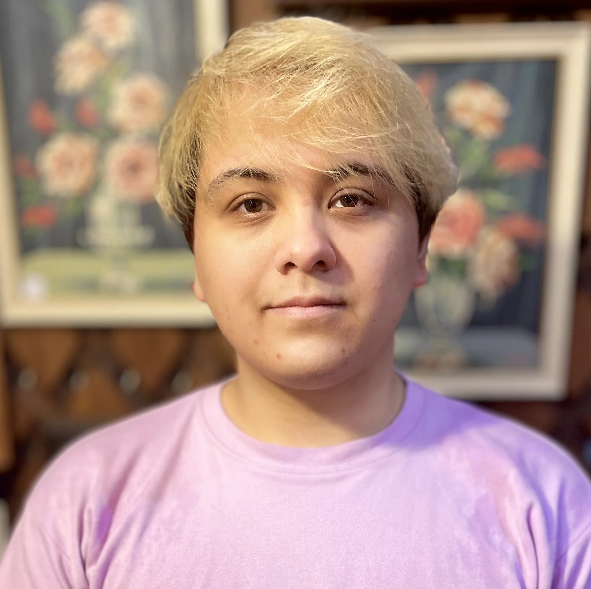 Brown man wearing a pink t-shirt looks at the camera. His hair is long on top and dyed blonde while the sides are short and dark. Behind him are framed paintings of flowers on stands.