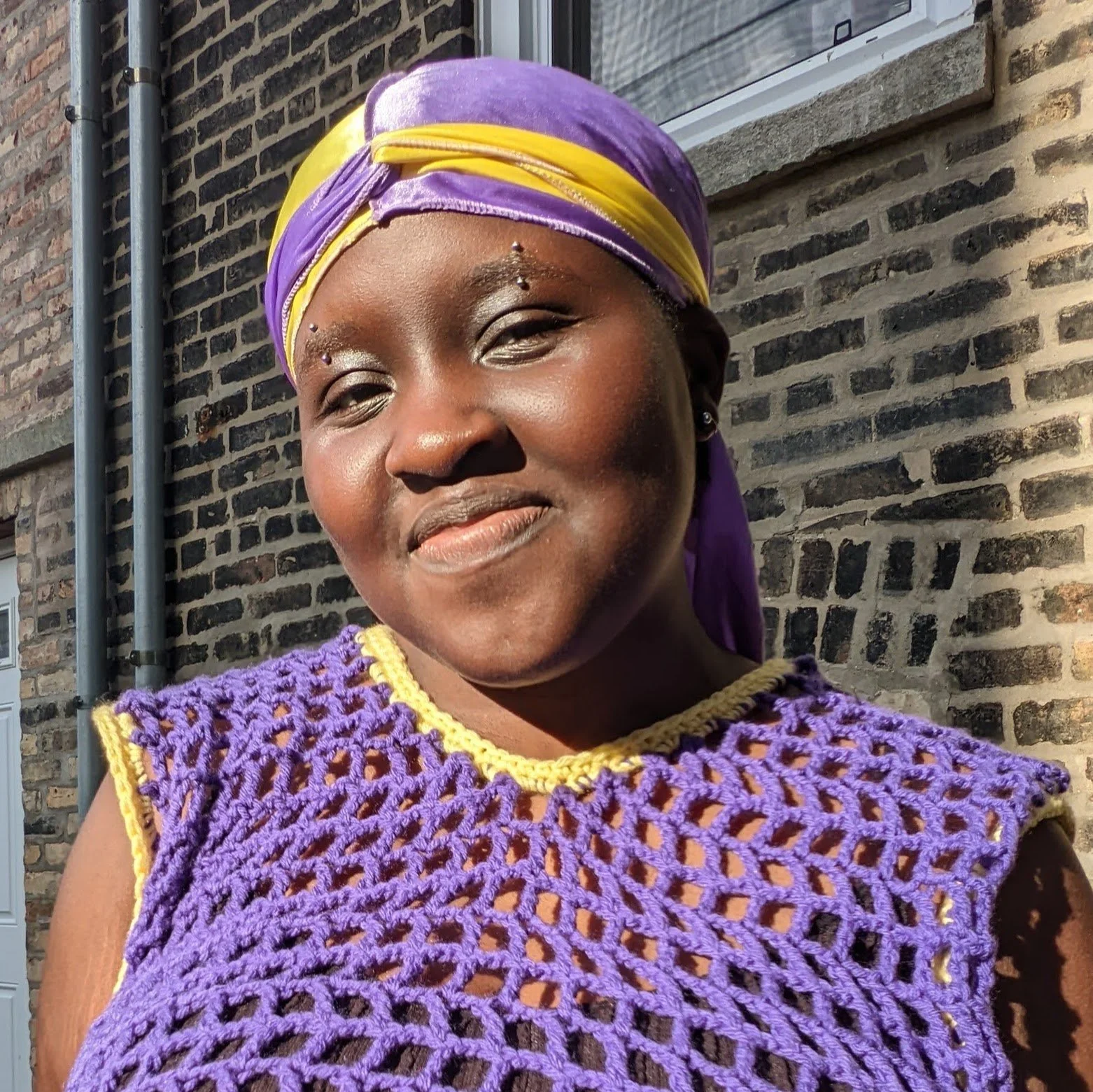 Black person with purple mesh top with yellow accents, smiling to camera.