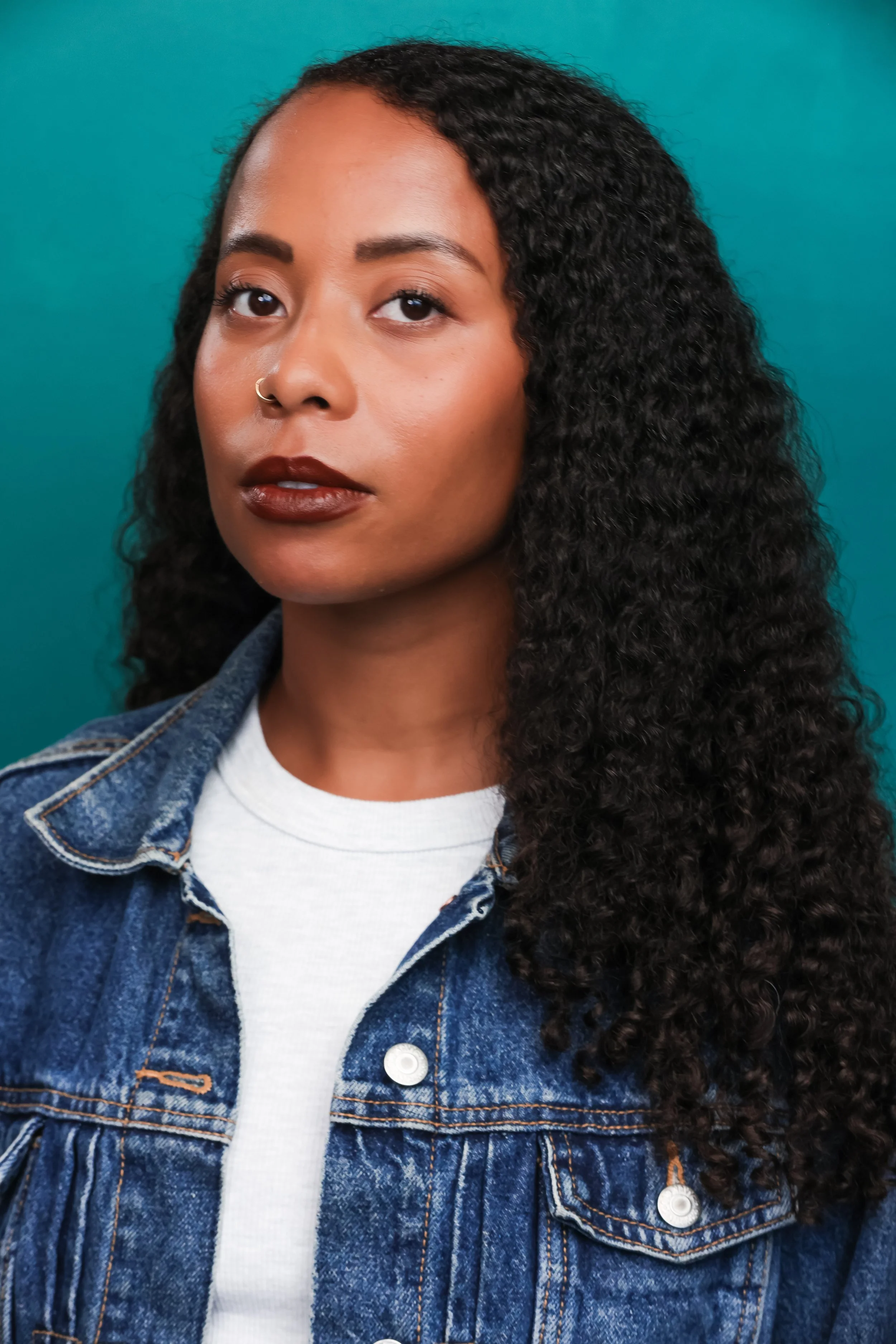 Black woman wearing white shirt with long curly dark hair, looking to camera.