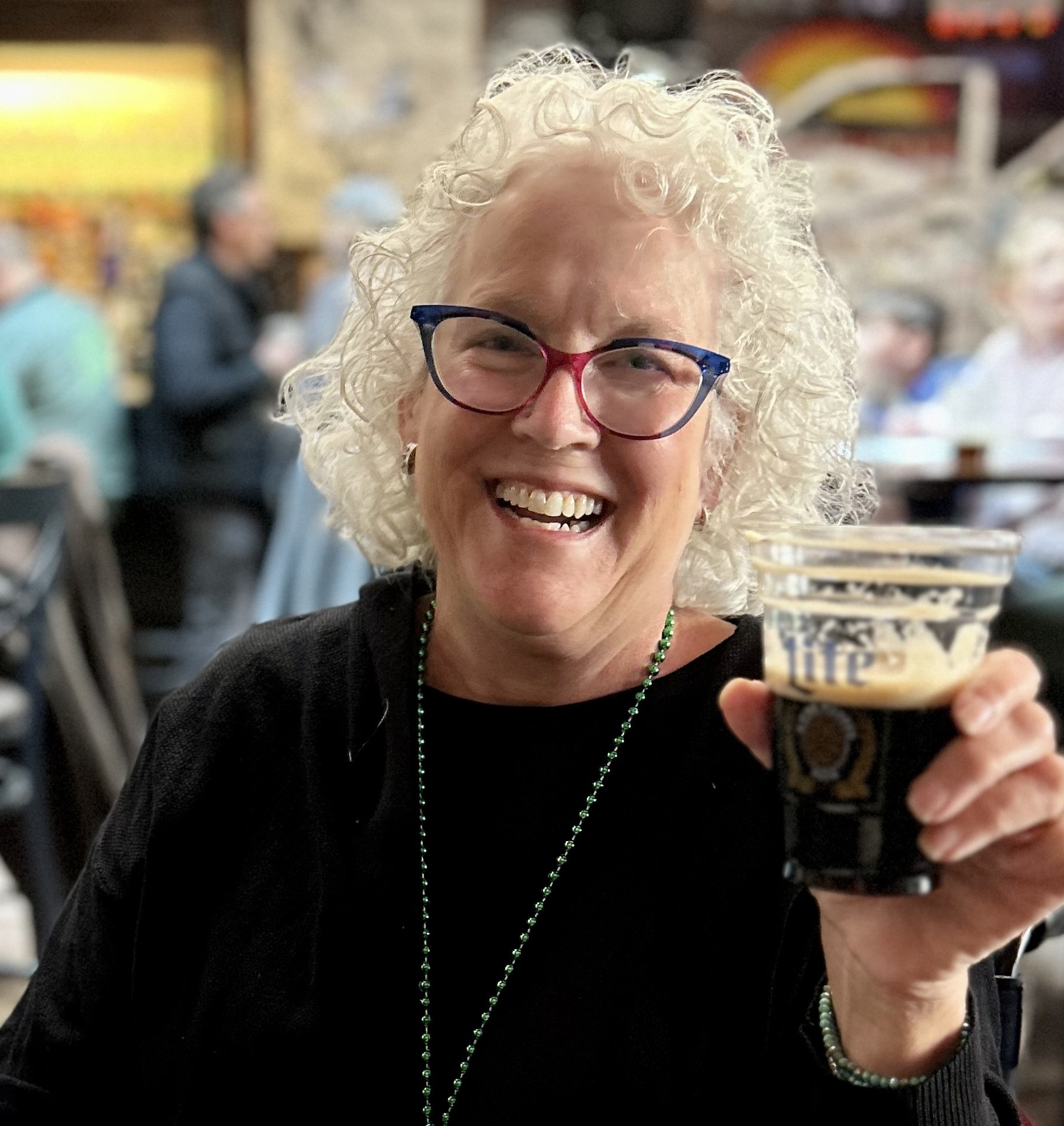 An older white woman with white curly hair smiles broadly while holding up a glass of dark beer as if to "cheers" to the viewer. She is wearing rainbow cat-eye glasses, a black sweater, and a long necklace.