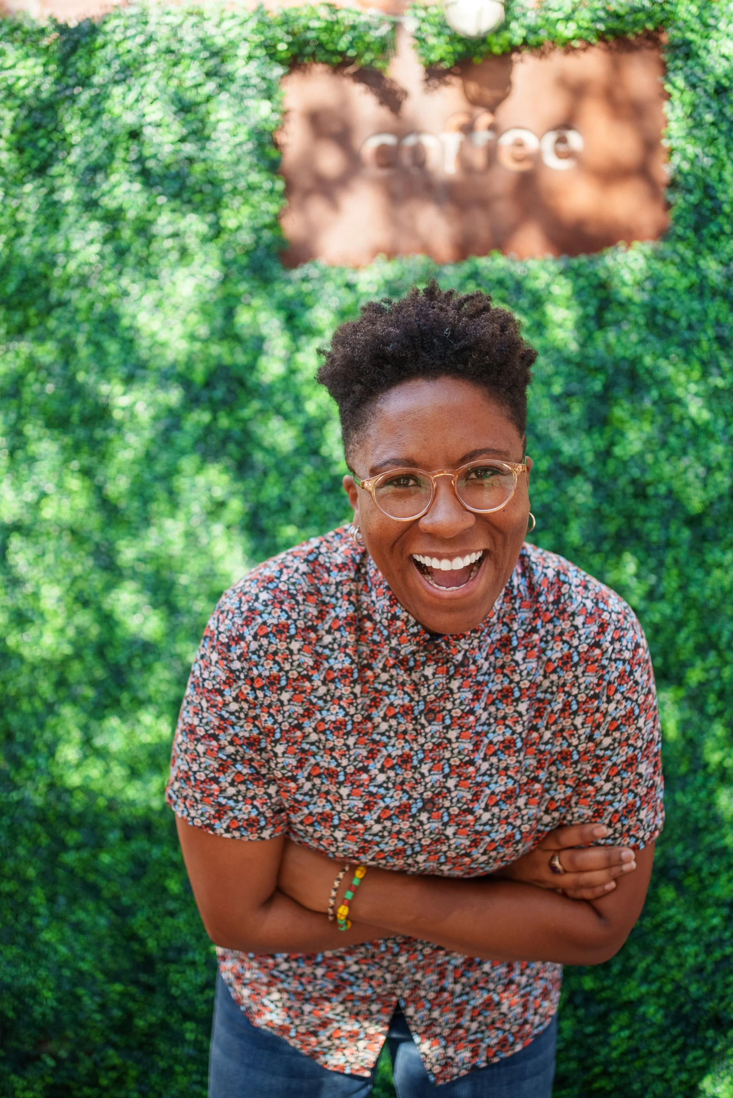 Black woman with short dark hair, wearing orange glasses, flower patterned shirt, smiling with mouth open to camera.