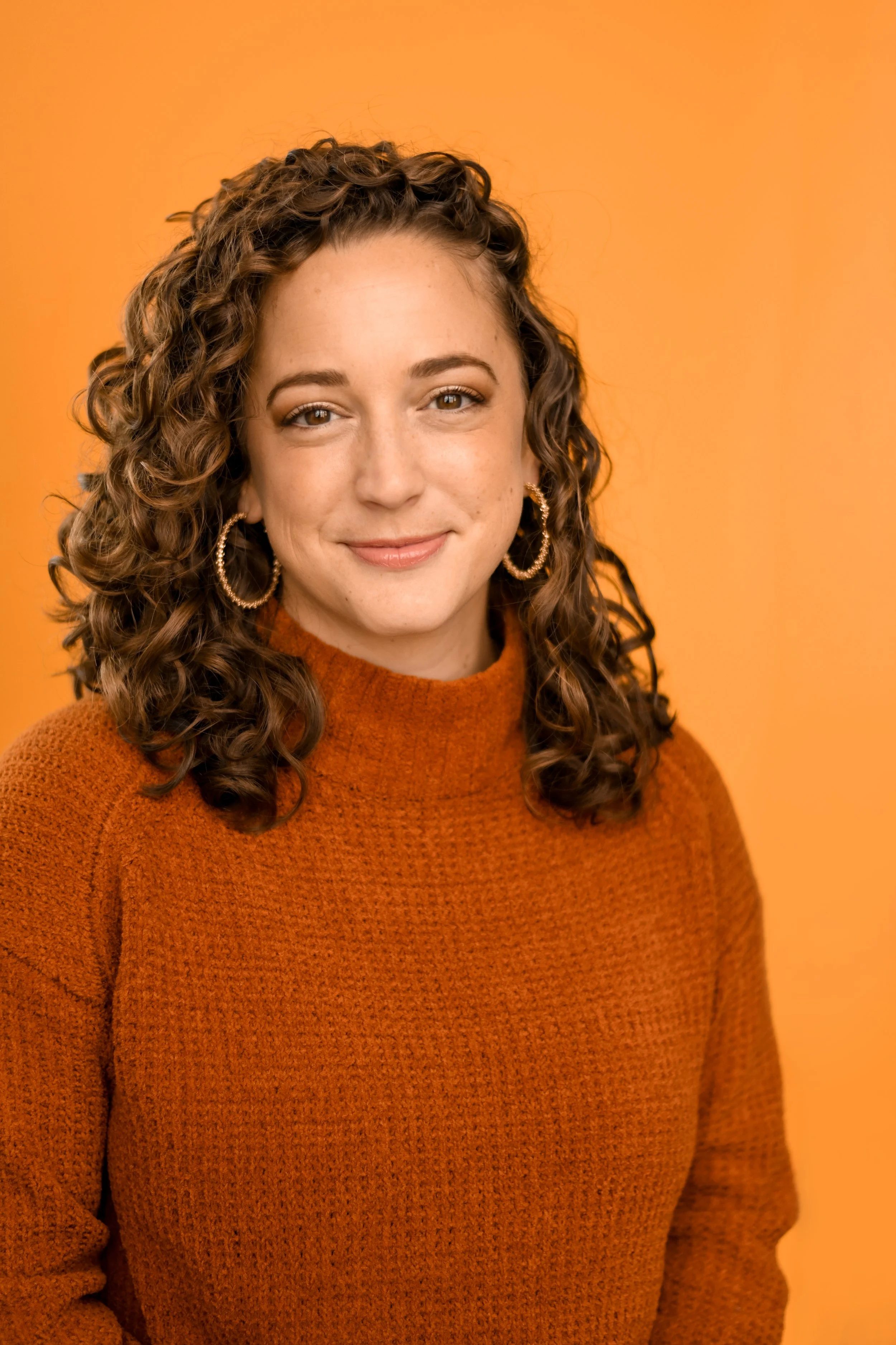 White person with curly brown hair and white skin, wearing hoops and an orange turtleneck.