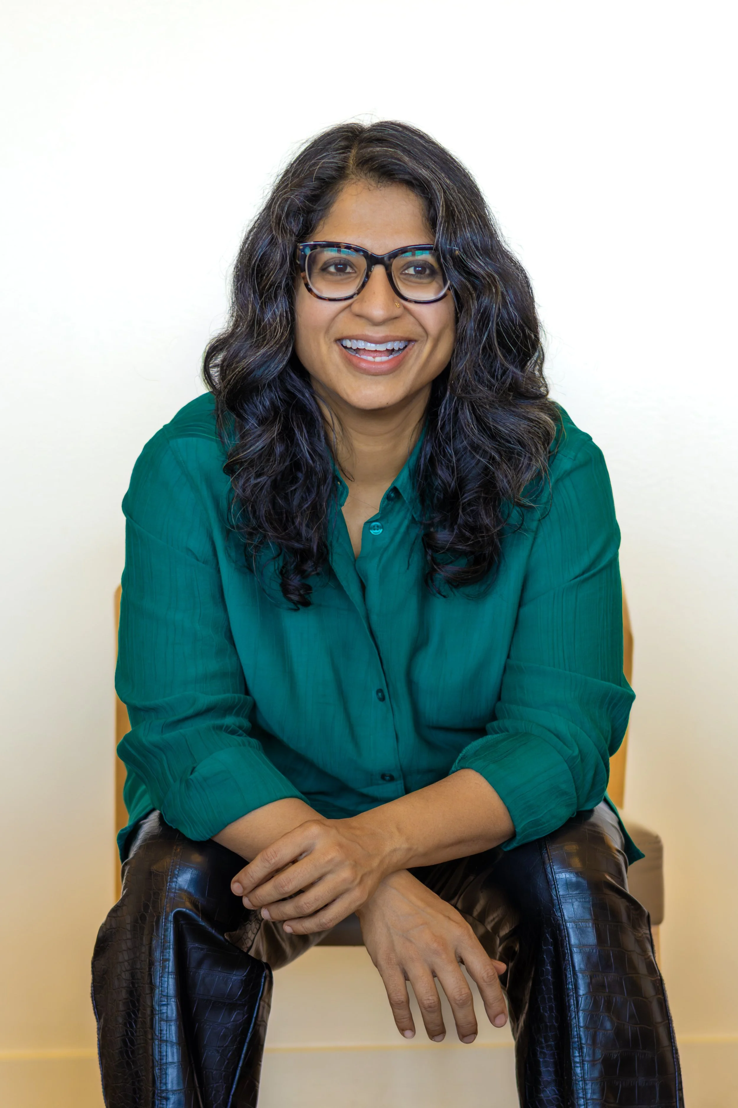 Woman with medium length dark hair, brown skin, glasses, and a teal button up shirt sits forward, smiling and looking at the camera.