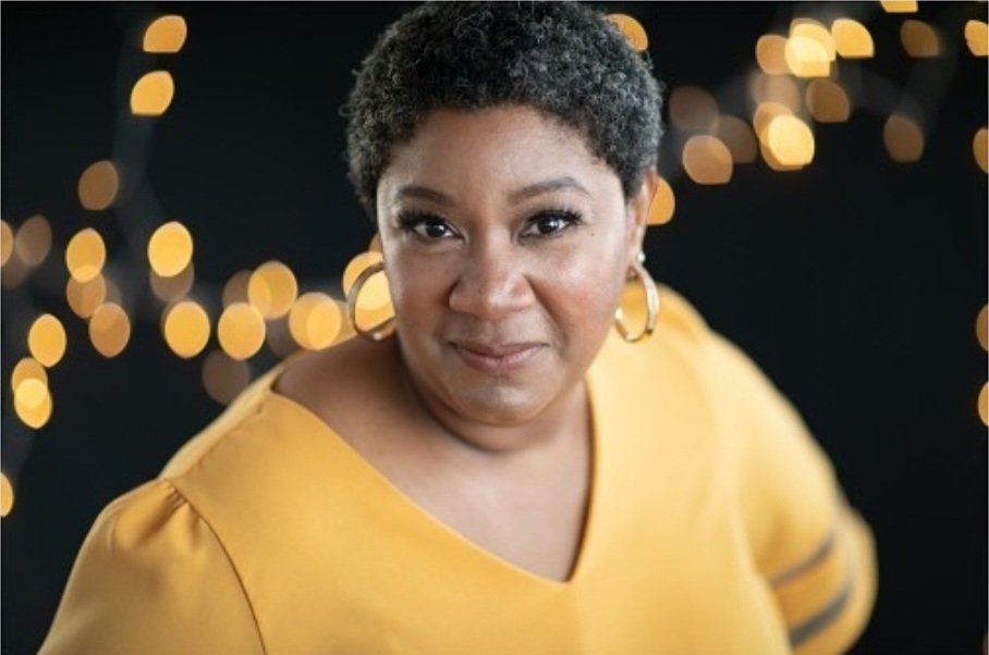 Black woman with short hair is wearing gold hoops and a yellow v-neck sweater. She is looking at the camera and the background is dark with blurry lights.