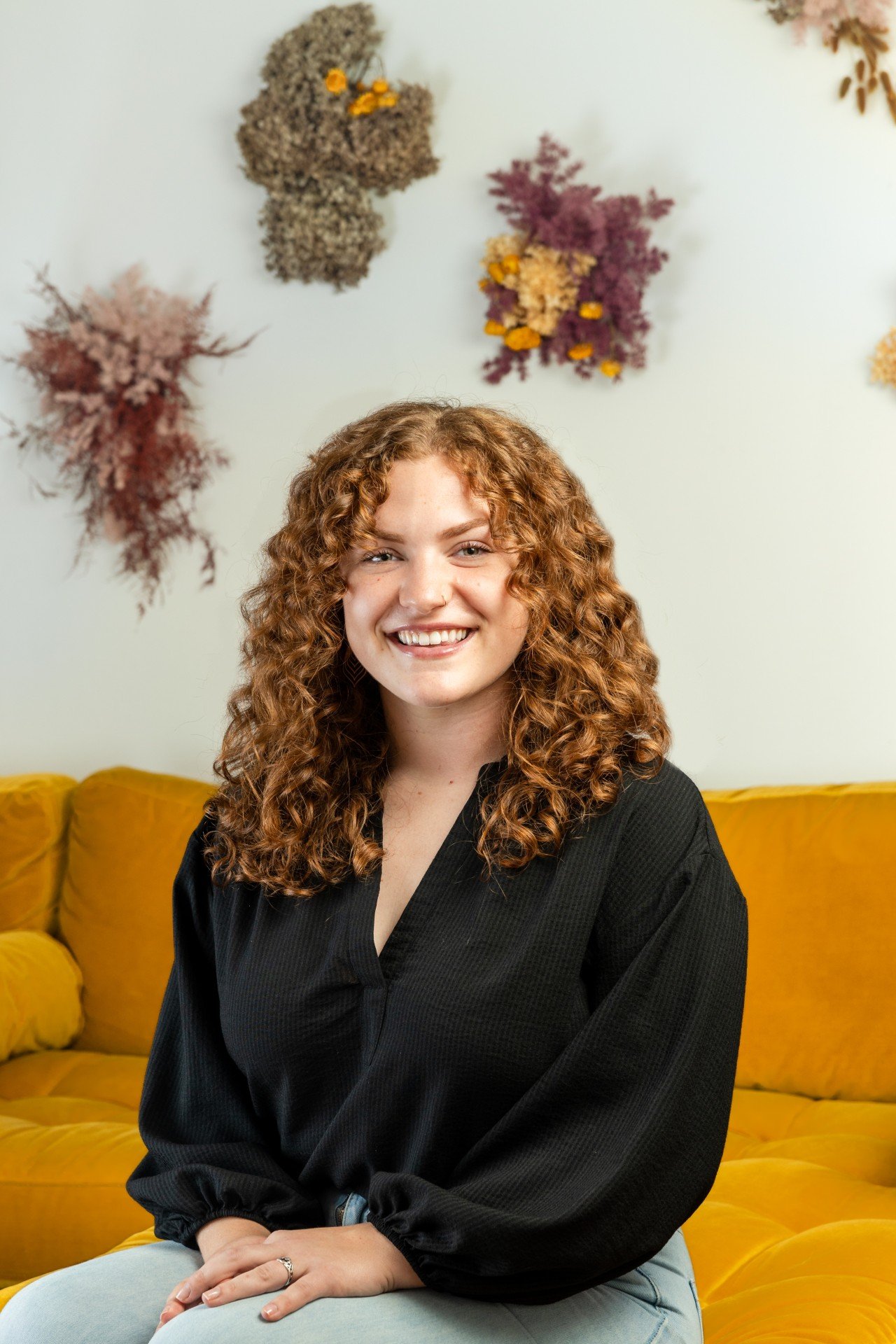 A white person sits on a mustard-yellow couch, smiling. They are wearing a black v-neck top.