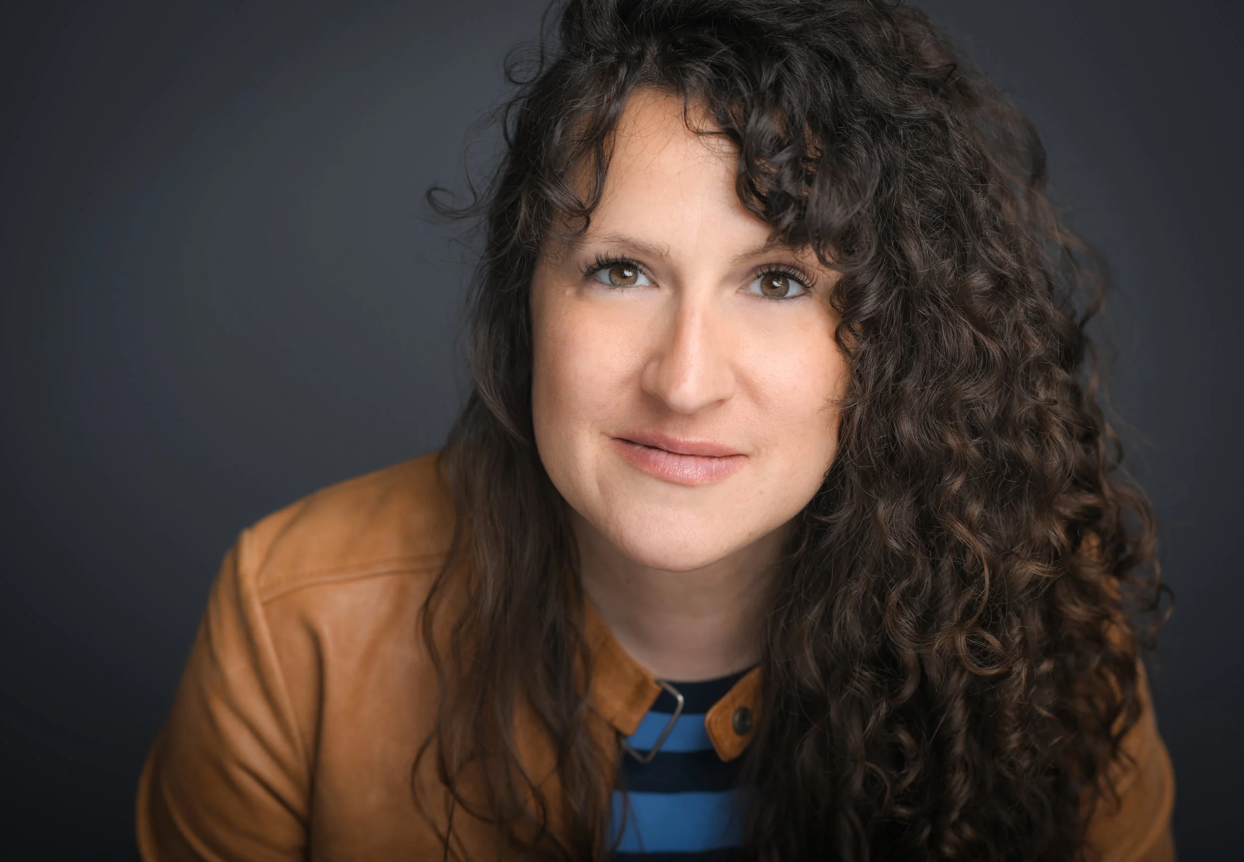 A white person with long, curly dark hair looks toward the camera, wearing a brown jacket over a striped shirt.