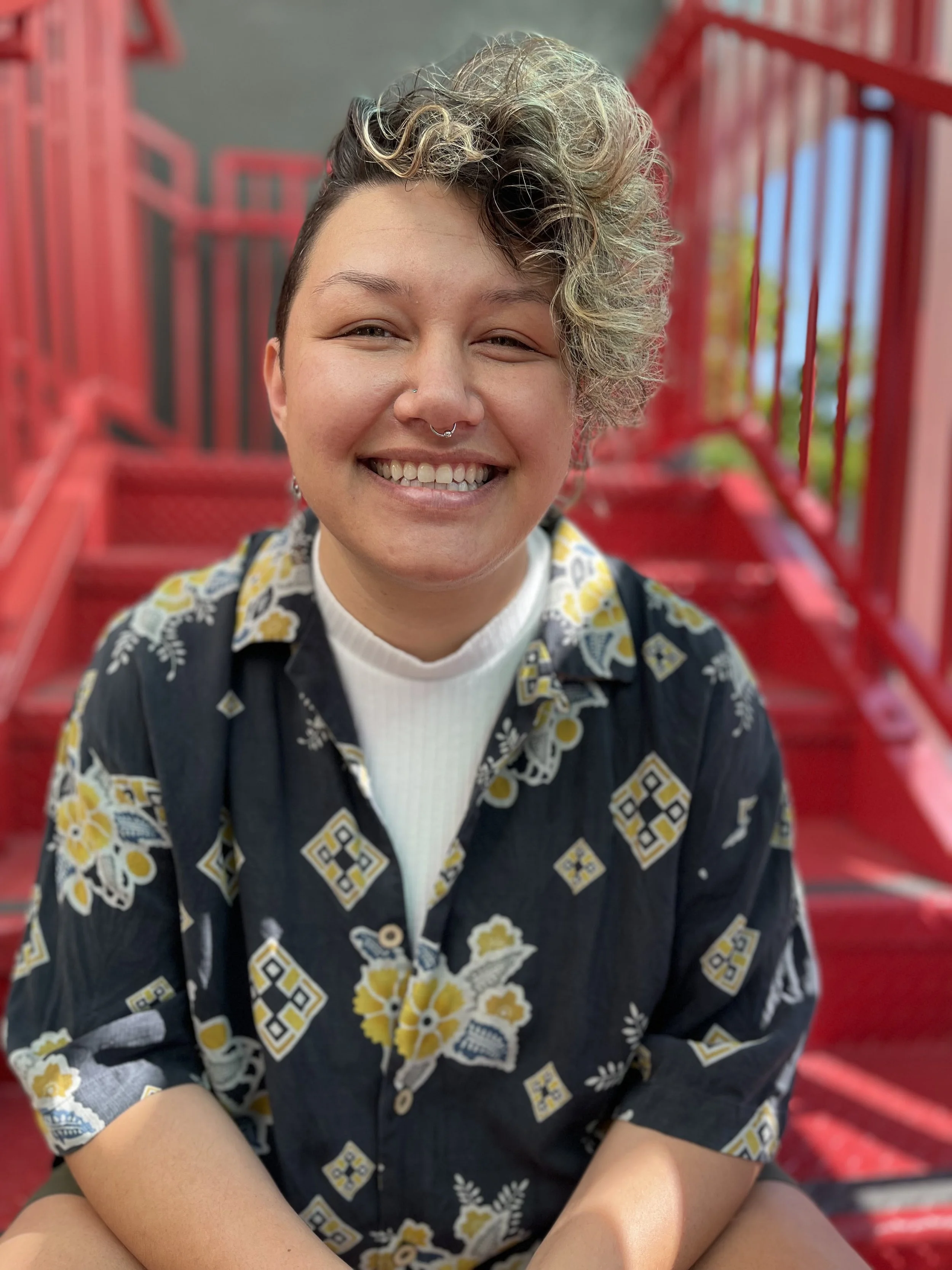 Person with short curly hair smiling to camera, wearing a white shirt with a patterned collar shirt.