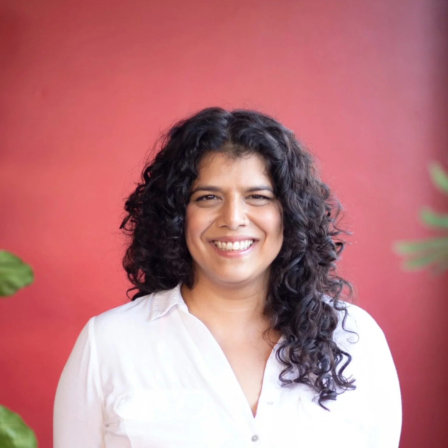 Brown woman with long curly dark hair stands against a red background wearing a white v-neck blouse and smiling