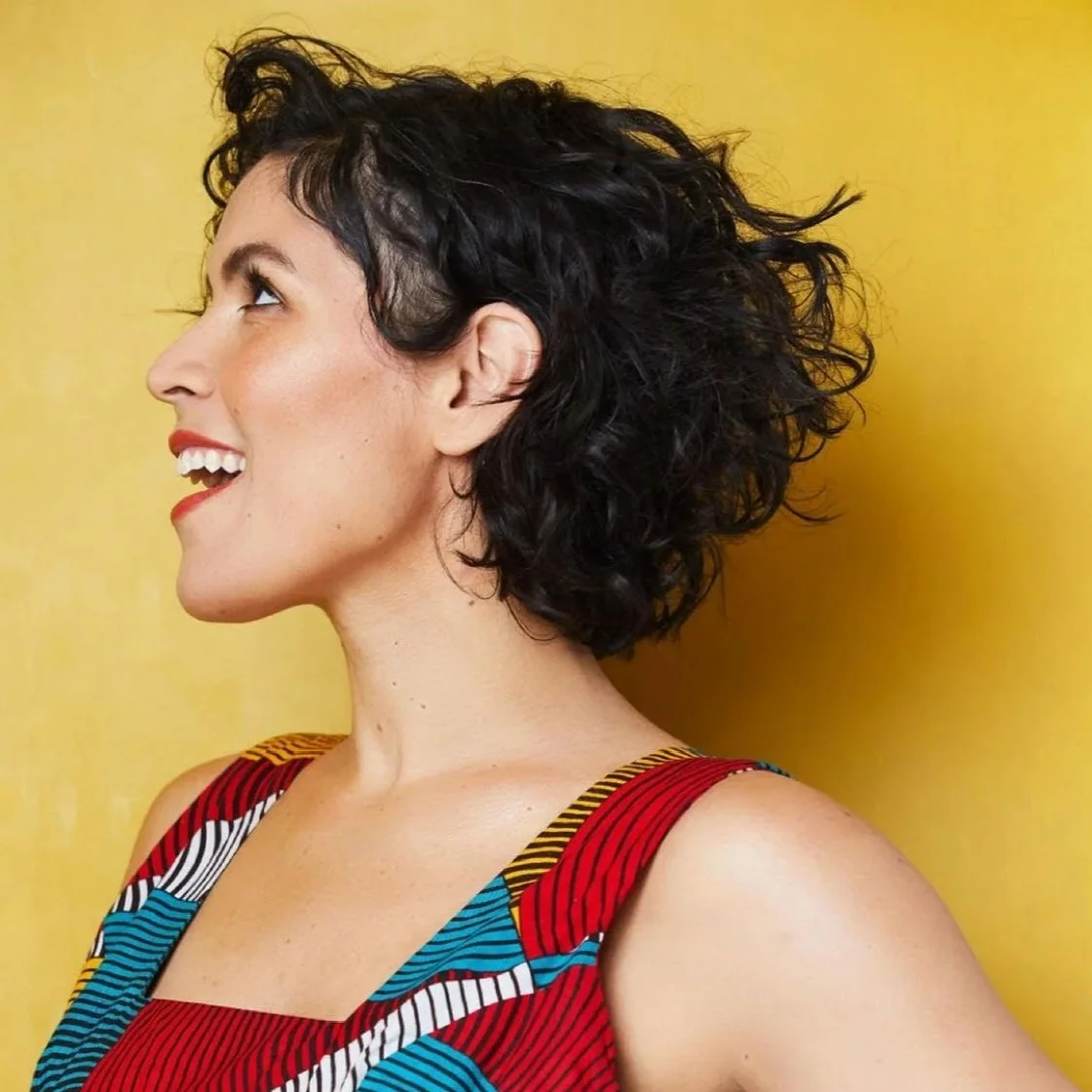 Woman with short curly dark hair, facing to the left of the camera smiling.