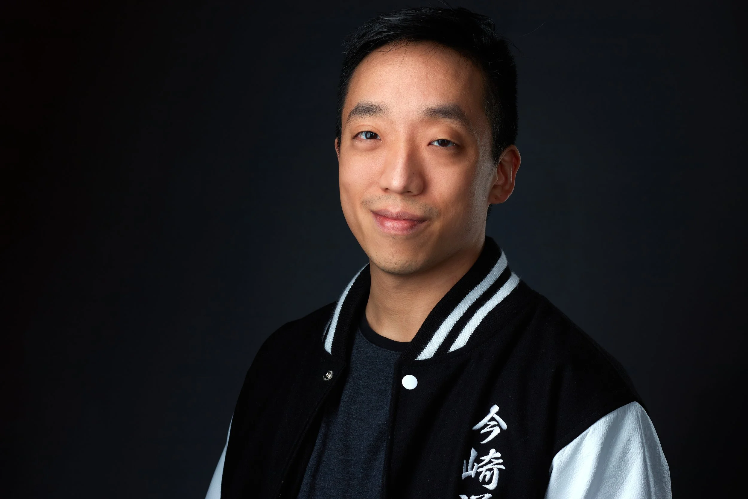 Vietnamese-American man smiles, closed mouthed, at the camera. He is wearing a dark black shirt and a black letterman-style jacket with white stripes on the collar.