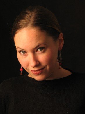 White woman looking to camera with dark hair pulled back, wearing dark shirt in front of a dark background.